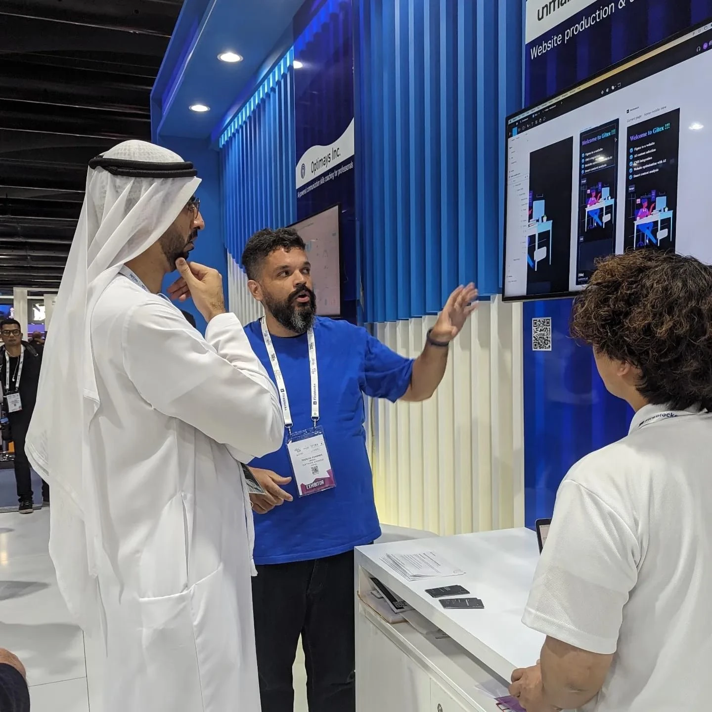 At an event booth, a presenter points to a product demo on a screen while two visitors listen.