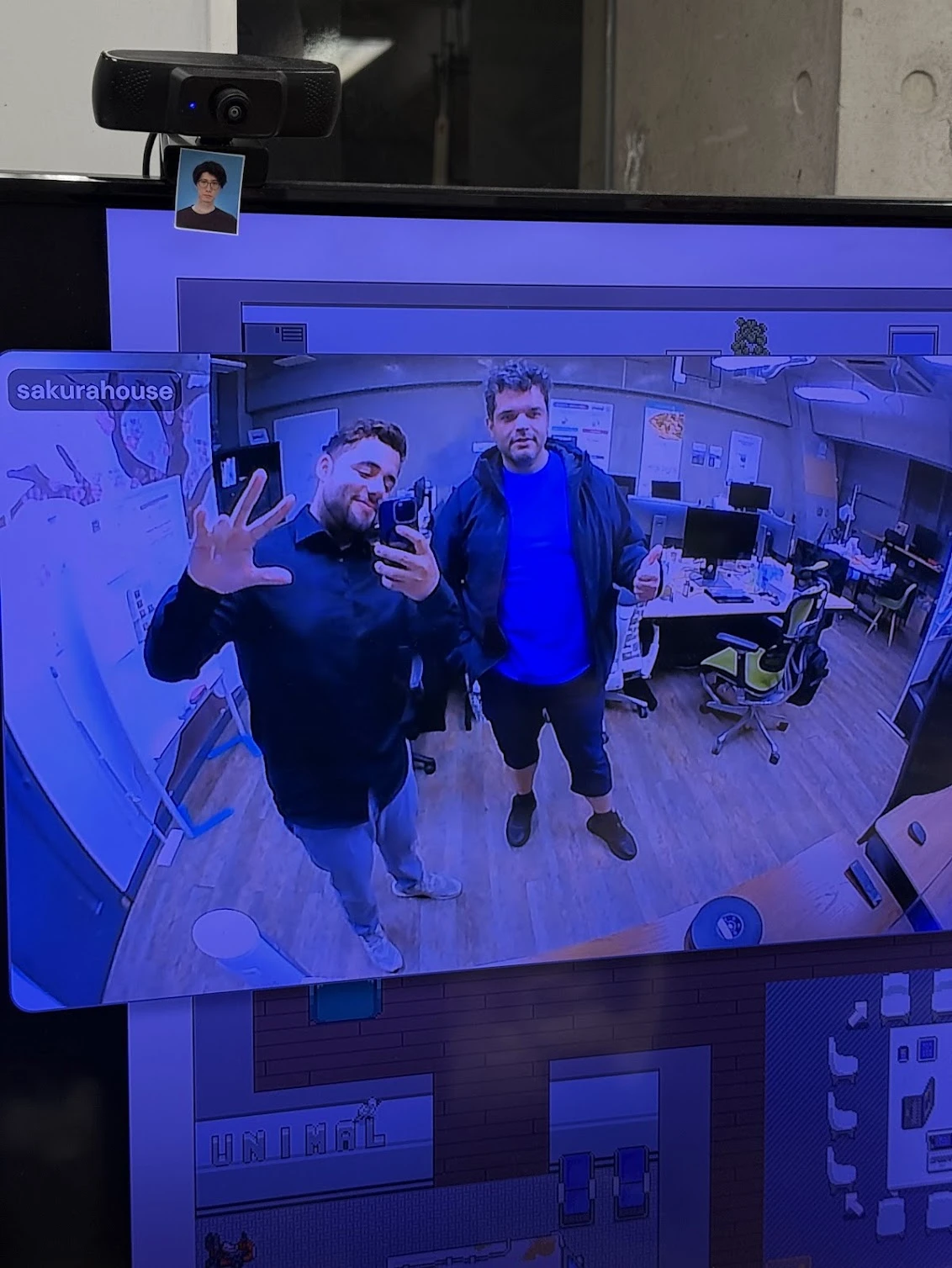 Two people wave at a webcam in an office, shown in a video tile on a large screen inside a pixel-art virtual office.